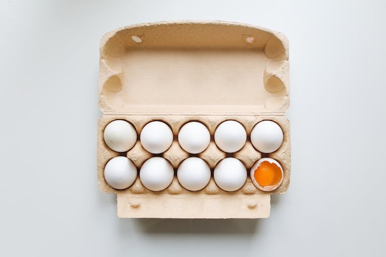 An open egg carton displaying eggs with one broken, revealing the yolk.